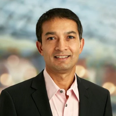 Smiling man in dark suit and light pink shirt standing against a blurred background.