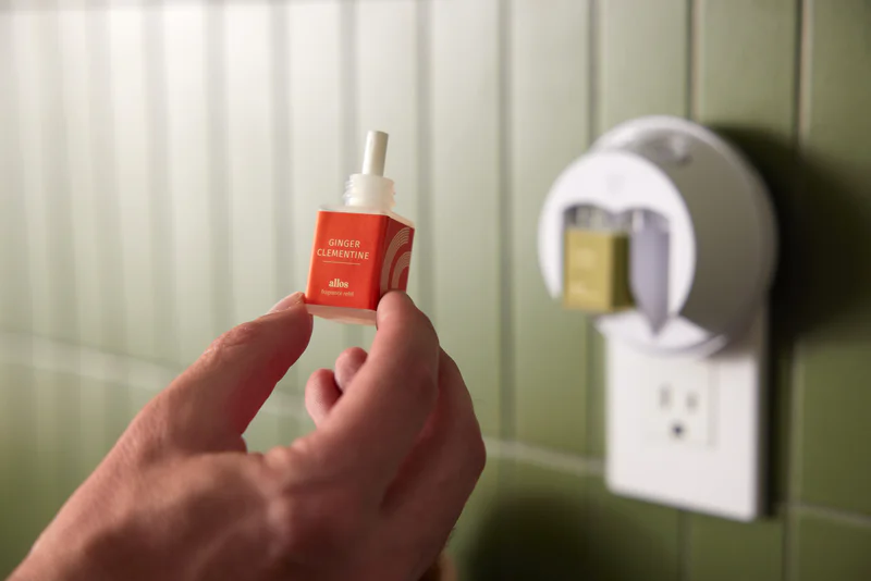 Person holding a ginger clementine fragrance refill next to a plug-in diffuser on a green-tiled wall. Person holding a ginger clementine fragrance refill next to a plug-in diffuser on a green-tiled wall.