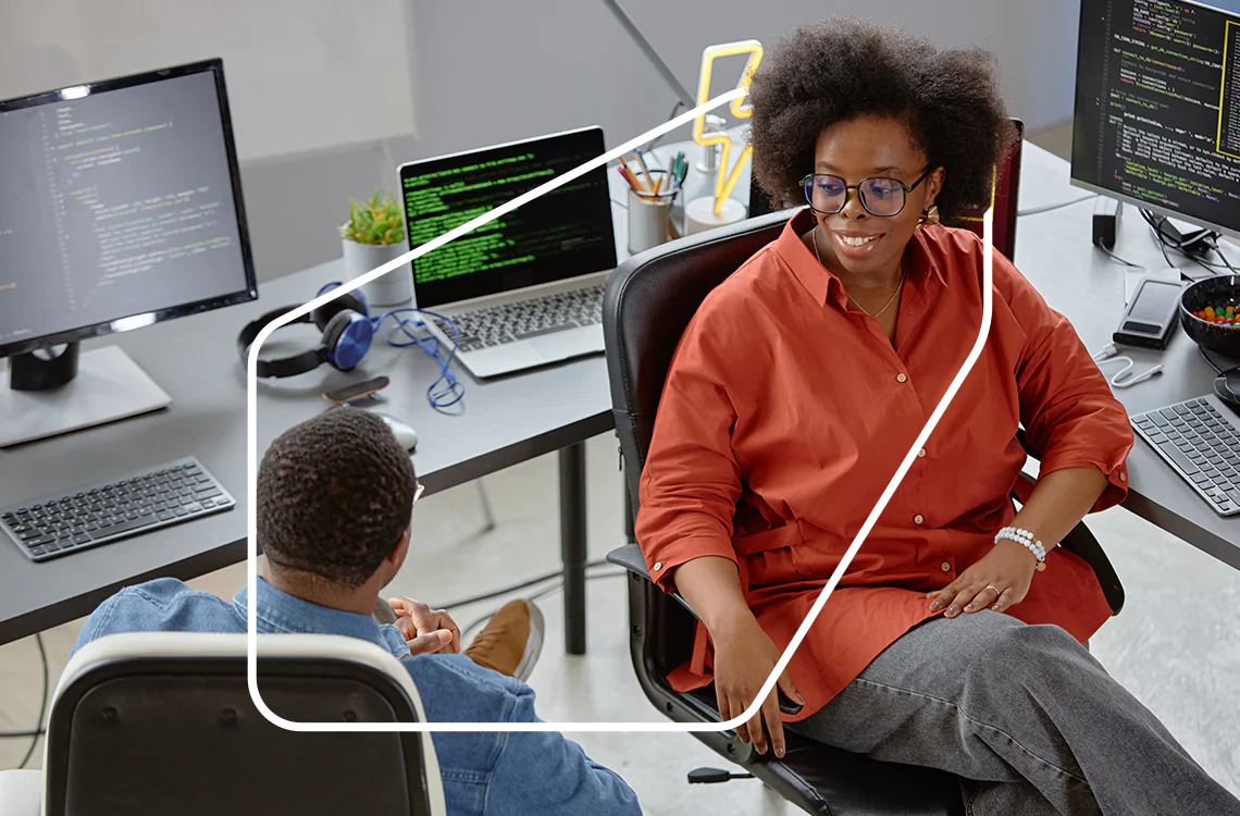 Two coworkers engage in discussion near desks with computers displaying code Two coworkers engage in discussion near desks with computers displaying code