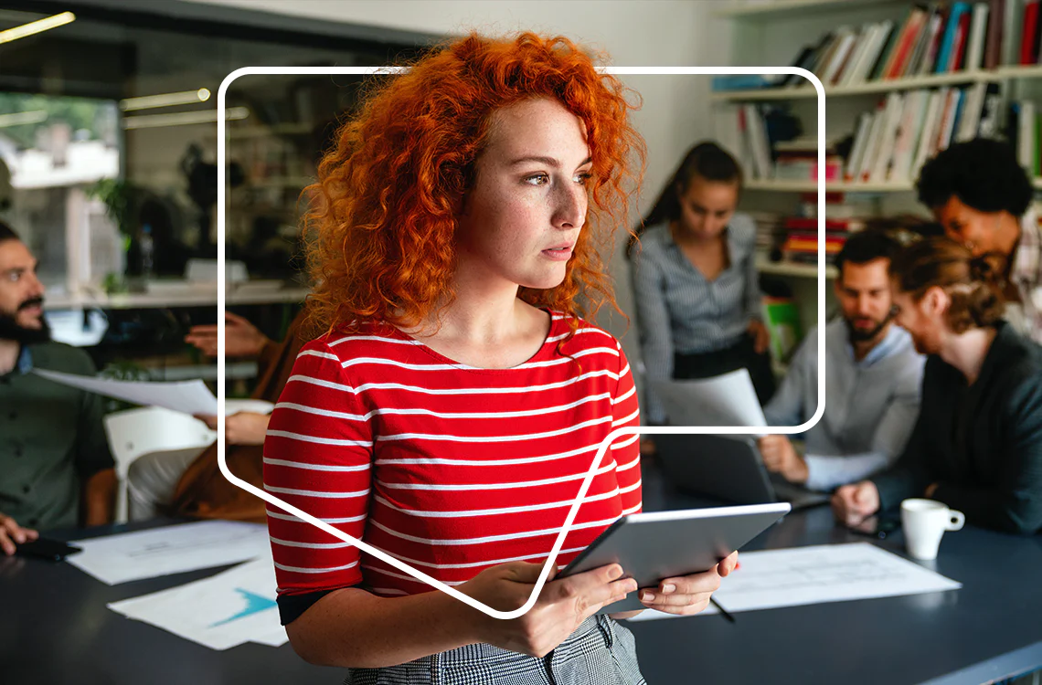 Woman with curly red hair and striped shirt uses tablet in an office with six other people working in the background. Woman with curly red hair and striped shirt uses tablet in an office with six other people working in the background.
