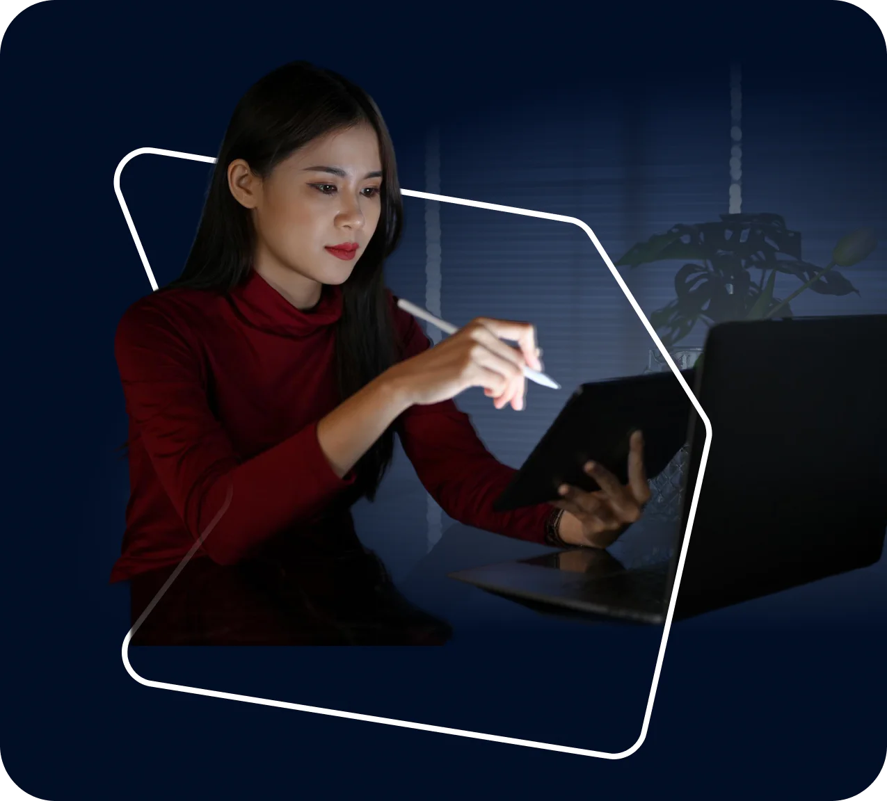 Woman using a stylus on a tablet at a desk with a laptop in a dimly lit room.