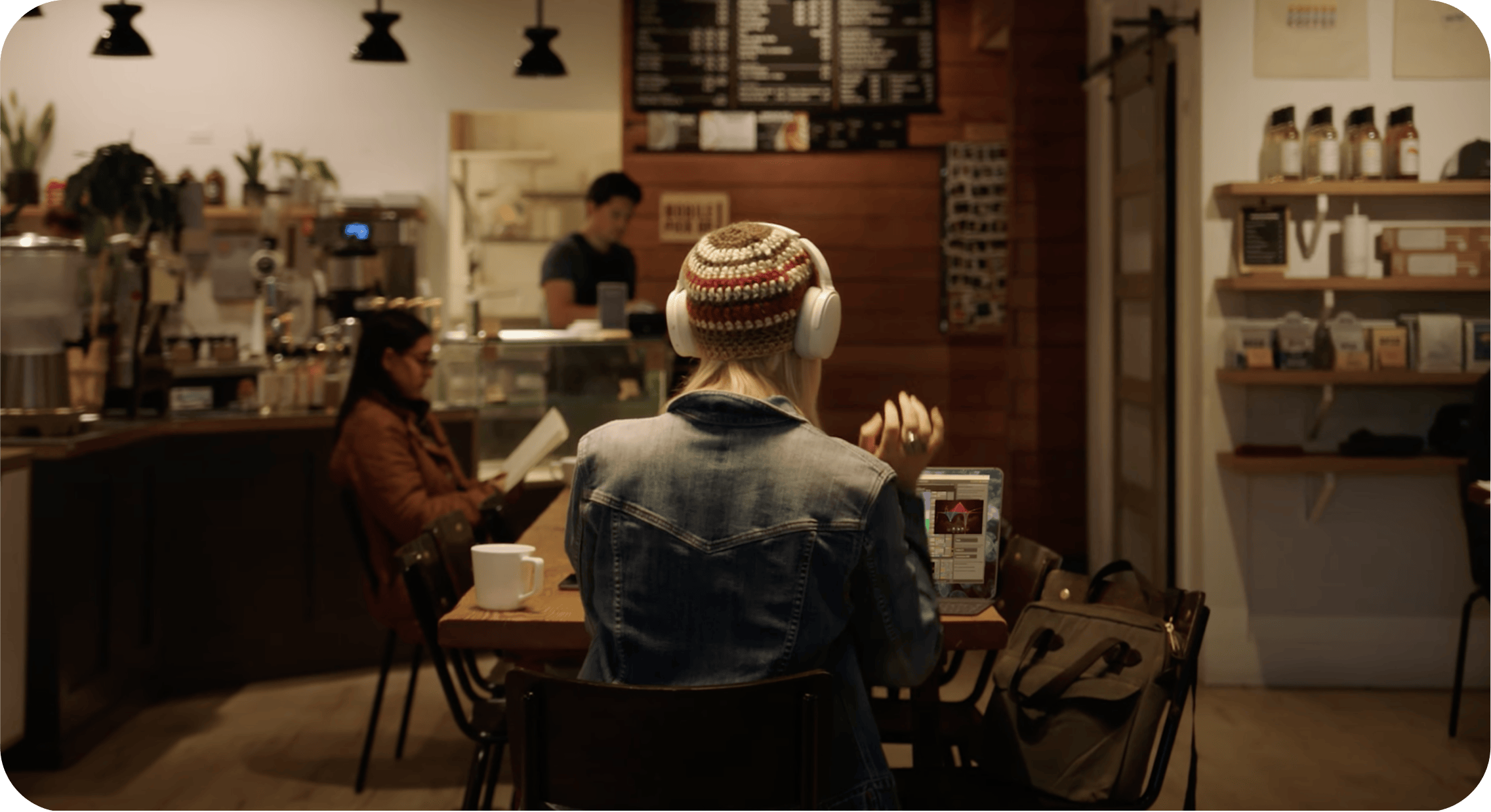 People sitting at a coffee shop, one wearing headphones and a beanie, with a barista in the background.